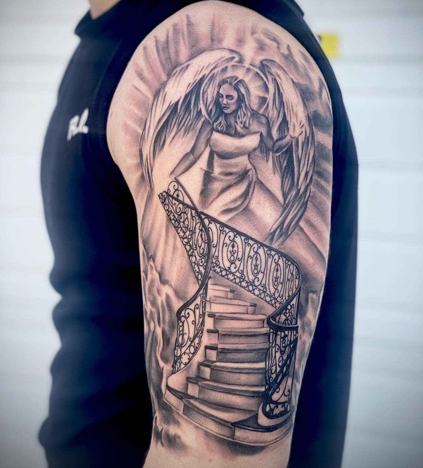 Tattoo of an angel with wings above ornate stairs on the upper arm